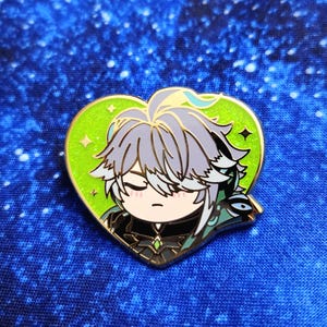 Può includere: Una spilla a forma di cuore in smalto con un personaggio dei cartoni animati con i capelli grigi e gli occhi chiusi. La spilla ha uno sfondo verde con dettagli dorati e un bordo dorato. Il personaggio indossa un abito nero.