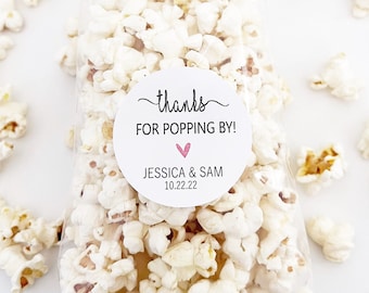 May include: A clear cellophane bag filled with white popcorn, with a round sticker that reads "thanks FOR POPPING BY!" with a pink heart, and the names "JESSICA & SAM" and the date "10.22.22". Scattered popcorn kernels surround the bag.