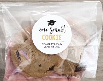 May include: A chocolate chip cookie in a clear plastic bag. A white circular sticker on the bag reads "one smart COOKIE" with a graduation cap graphic and "CONGRATS JOHN CLASS OF 2022" in black and gold.