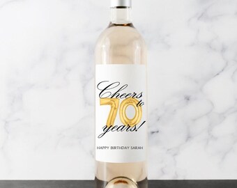 Może przedstawiać: Przezroczysta butelka wina ze srebrną zakrętką i białą etykietą. Na etykiecie napis "Cheers to 70 years!" czarnym pismem ze złotym balonem "70". Poniżej napis "Happy Birthday Sarah".