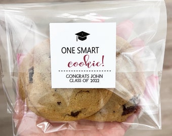 May include: Two chocolate chip cookies in a clear cellophane bag. A white label on the bag reads "ONE SMART cookie!" in black and burgundy text, with a graduation cap icon. Below the text is "CONGRATS JOHN CLASS OF 2022".