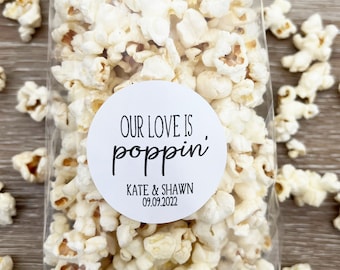 May include: A clear cellophane bag filled with popcorn. A white circular sticker on the bag reads "OUR LOVE IS poppin'" with the names "KATE & SHAWN" and the date "09.09.2022" in black text. Scattered popcorn kernels are visible around the bag.