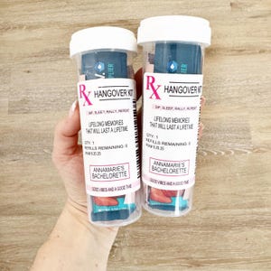 Może przedstawiać: Dwie przezroczyste plastikowe tuby z białymi wieczkami, oznaczone jako "Rx Hangover Kit". Tuby zawierają niebieską i białą zawartość oraz tekst "Lifelong Memories That Will Last a Lifetime". Na dolnej etykiecie napisano "Annamarie's Bachelorette" i "Good Vibes and a Good Time."