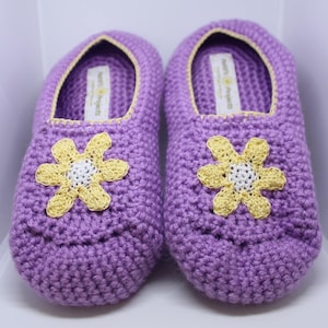 Peut inclure: Une paire de pantoufles en crochet violet avec des fleurs jaunes. Les pantoufles ont une bordure jaune clair autour des bords et le texte "Knit Project" est visible à l'intérieur des pantoufles.