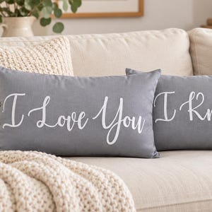 May include: Two rectangular grey throw pillows featuring white cursive text. The left pillow displays the phrase I Love You, while the right pillow displays the phrase I Know. Both pillows have a smooth fabric finish and are positioned side by side.
