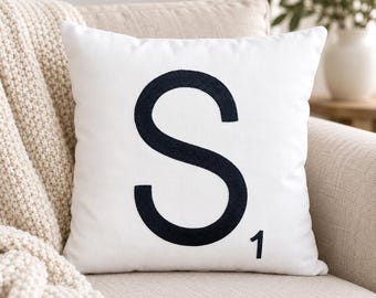 May include: A square white throw pillow featuring a large, centered, dark navy blue embroidered letter S. The letter is styled in a bold, sans-serif font, and a small number one is embroidered in the same dark navy blue color in the bottom right corner.