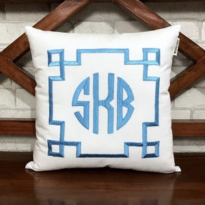 May include: White square cushion featuring a light blue monogram and geometric border design. The monogram displays the letters SKB in a circular arrangement, embroidered in a soft blue. The cushion is placed on a dark wooden surface.