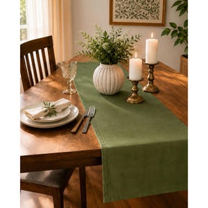 Op de afbeelding: Een effen olijfgroene tafelloper op een houten eettafel. Het middelpunt bestaat uit een witte geribbelde keramische vaas met groen blad, twee koperen kandelaars met witte stompkaarsen, een glazen beker en een wit bord.