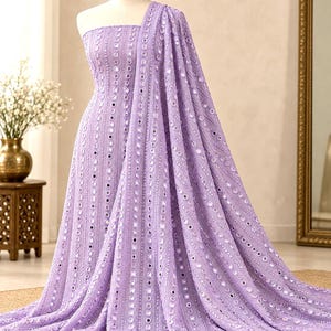 Puede incluir: Un vestido palabra de honor de color lavanda con filas verticales de intrincados bordados y pequeños espejos circulares. La tela está drapeada para crear una silueta fluida, con una pieza de tela larga a juego que cae desde el hombro al suelo.