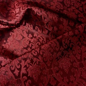 May include: Close-up of a deep red fabric with a damask pattern. The fabric has a subtle sheen and a textured surface.