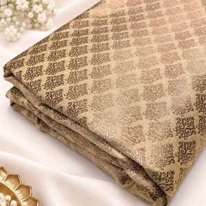 Puede incluir: Una pieza de tela doblada en color beige claro con un patrón de damasco marrón oscuro repetitivo. El tejido tiene un brillo sutil y está dispuesto en varias capas, mostrando el intrincado diseño tejido en toda la superficie.
