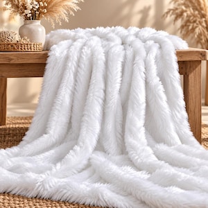 May include: A plush white faux fur throw blanket with a long, shaggy texture. The blanket is draped over a wooden surface, showcasing its thick, soft pile and bright white colour. The material appears dense and fluffy, covering the surface area.