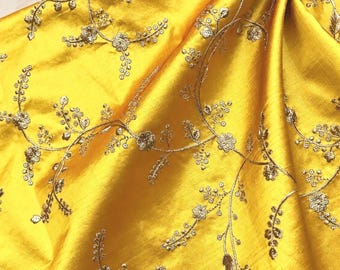 Puede incluir: Una tela de color amarillo vibrante con un intrincado bordado de hilo dorado en un delicado patrón de enredadera floral. El material tiene un brillo sutil y una textura de cuadrícula fina, con costuras metálicas en relieve.