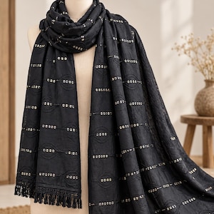 May include: A black scarf featuring horizontal rows of gold-toned sequins and embroidered circular patterns. The fabric has a textured appearance and the edges are finished with a decorative lace trim and a fringe of black tassels along the bottom.