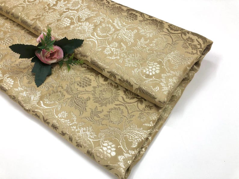 May include: Folded gold fabric with a floral pattern. The fabric has a shiny finish and is decorated with a small pink rose and green leaves. The fabric is likely used for clothing or home decor.
