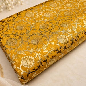 May include: A folded piece of mustard yellow fabric featuring an intricate, repeating metallic gold floral and vine pattern. The textile has a dense, woven texture with a shimmering finish, showcasing detailed botanical motifs across the surface.