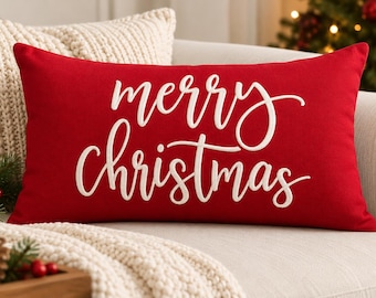 May include: A rectangular red throw pillow featuring the white embroidered text merry christmas in a cursive script font. The pillow has a smooth fabric surface and is positioned horizontally, displaying the holiday greeting clearly across the center.