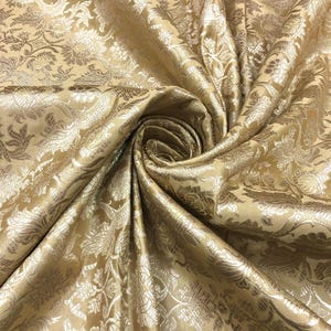 May include: Close-up of a golden-colored fabric with a floral pattern. The fabric is folded and draped, creating a swirling effect. The pattern features detailed floral and leaf designs in a lighter gold hue, adding depth and texture to the material.