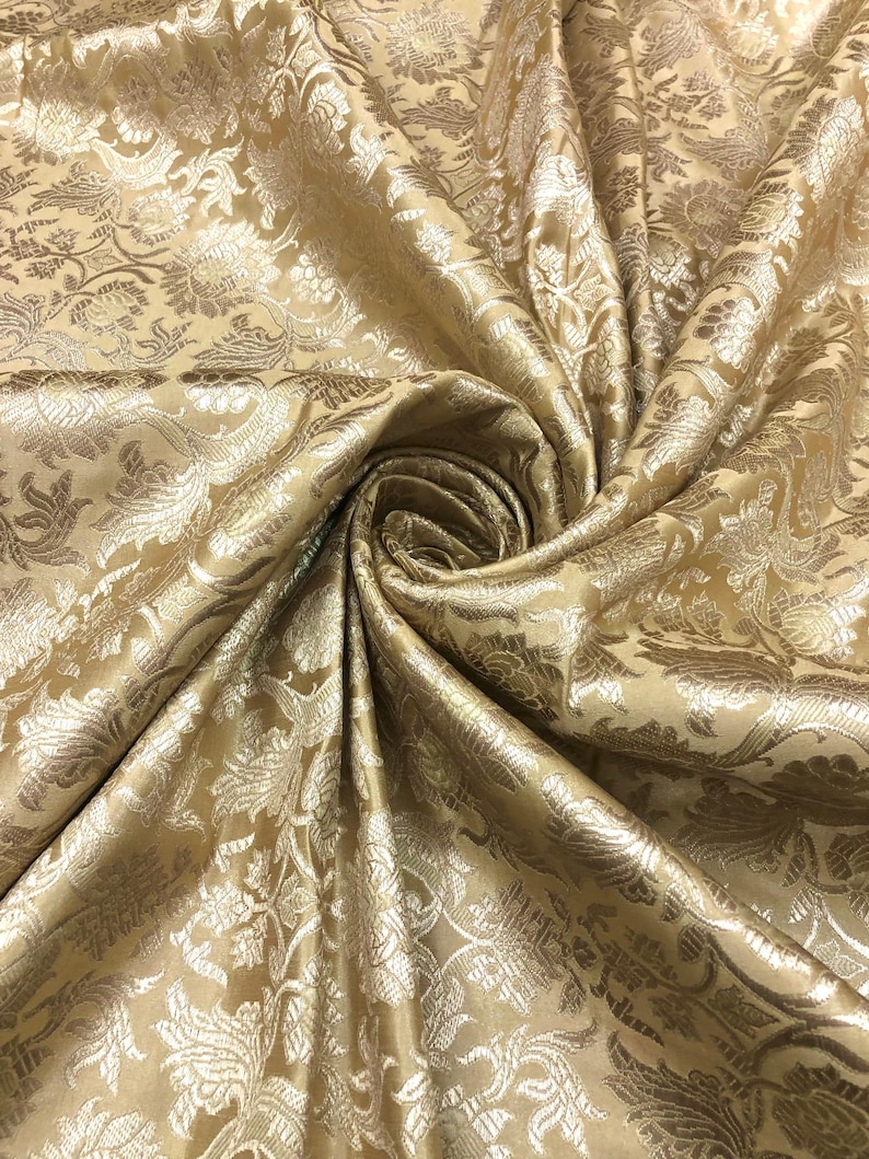 May include: Close-up of a golden-colored fabric with a floral pattern. The fabric is folded and draped, creating a swirling effect. The pattern features detailed floral and leaf designs in a lighter gold hue, adding depth and texture to the material.