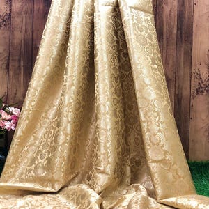 May include: A draped, golden-colored fabric with a detailed floral pattern. The material has a subtle sheen, and the folds create shadows that highlight the intricate design. The fabric appears to be heavy and luxurious, suitable for formal wear or home decor.