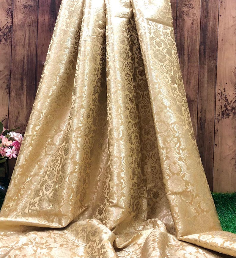 May include: A draped, golden-colored fabric with a detailed floral pattern. The material has a subtle sheen, and the folds create shadows that highlight the intricate design. The fabric appears to be heavy and luxurious, suitable for formal wear or home decor.