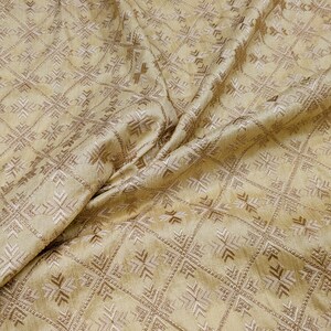 Peut inclure: Un tissu doré clair présentant un motif géométrique répétitif de lignes brodées blanches et dorées métalliques. Le textile affiche un design en grille structuré avec des motifs complexes en forme de croix centrés dans chaque section carrée.