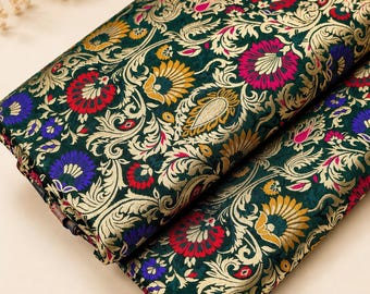 Puede incluir: Una pieza de tela verde oscuro doblada que presenta un intrincado patrón tejido de motivos florales y de enredaderas en dorado, rojo, azul y amarillo. El tejido muestra un diseño denso y ornamentado con hilos de color metálico.
