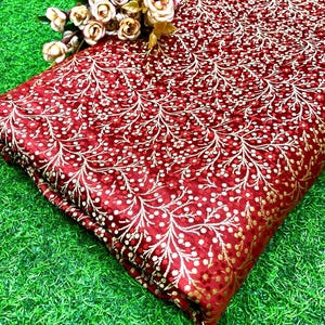 Può includere: Un pezzo di tessuto rosso piegato con un motivo floreale dorato. Il tessuto ha un aspetto lucido e strutturato. Un piccolo bouquet di rose color crema poggia sul tessuto. Il tessuto è su una superficie verde.