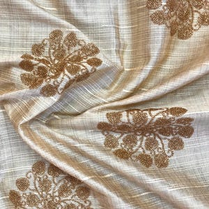 Puede incluir: Tela beige con un sutil patrón a rayas y diseños florales bordados en color dorado.