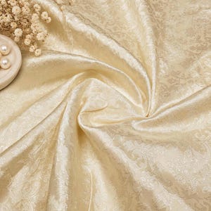 May include: A cream-colored fabric featuring a tonal damask pattern with a subtle sheen. The material is gathered in the center, creating soft folds and drapes. A small cluster of dried white flowers and two white pearls are visible.