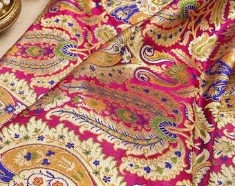 Puede incluir: Una tela fucsia vibrante con un intrincado patrón tejido de grandes motivos de cachemira en dorado, azul y verde. El textil muestra un diseño denso y ornamentado con hilos de oro metálico, terminado con un borde detallado de rayas horizontales.