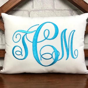 May include: A rectangular white cushion featuring a large, embroidered monogram in a vibrant turquoise colour. The monogram has three stylized letters with elegant, swirling details, creating a personalized decorative accent.