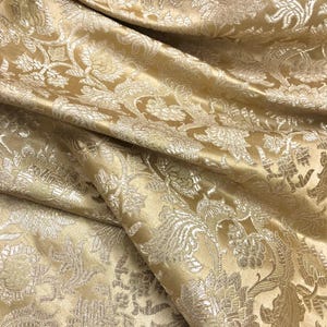 May include: Close-up of a golden-yellow fabric with a detailed floral pattern. The fabric has a satin sheen and is folded, creating shadows and highlights. The design features stylized flowers and leaves.