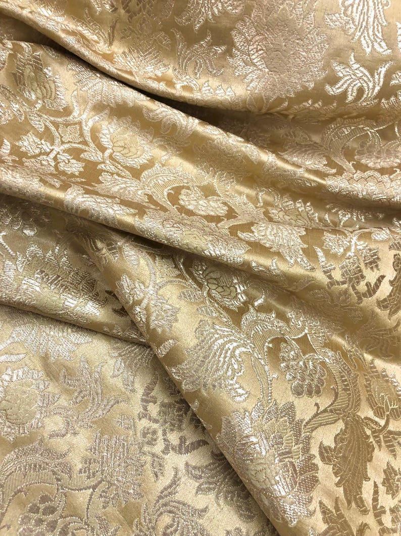 May include: Close-up of a golden-yellow fabric with a detailed floral pattern. The fabric has a satin sheen and is folded, creating shadows and highlights. The design features stylized flowers and leaves.