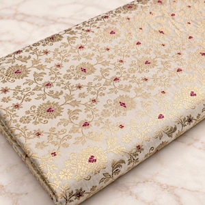 Puede incluir: Una pieza de tela beige claro doblada que presenta un intrincado patrón tejido de enredaderas florales en oro metálico y pequeños acentos magenta. El textil muestra un diseño botánico denso y repetitivo en toda su superficie visible.