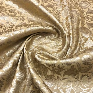 May include: Close-up of a golden fabric with a floral pattern. The fabric has a shiny, textured surface with intricate details in a lighter gold color. The folds and drapes of the fabric create shadows and highlights, enhancing the visual texture.
