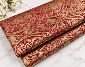 Könnte beinhalten: Ein gefaltetes Stück Stoff mit tiefrotem Hintergrund und einem komplizierten, sich wiederholenden goldenen Paisley- und Blumenmuster. Das Textil hat eine gewebte Struktur mit metallischen Goldfäden, die ein detailliertes, verziertes Design bilden.