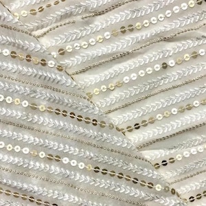 Op de afbeelding: Witte stof met gouden pailletten die in horizontale rijen zijn gerangschikt. De pailletten zijn genaaid in een patroon van kleine bladeren.