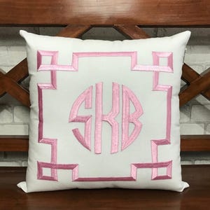 May include: White square cushion with a pink embroidered geometric design and monogram. The cushion features a central monogram with the letters "SKB" in a pink, stylized font. The design is set against a white background.