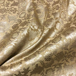 May include: Close-up of a golden fabric with a floral pattern. The fabric has a shiny, textured surface with detailed designs of flowers and leaves. The folds in the fabric create shadows and highlights, enhancing the intricate details of the pattern.