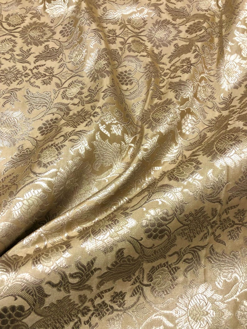 May include: Close-up of a golden fabric with a floral pattern. The fabric has a shiny, textured surface with detailed designs of flowers and leaves. The folds in the fabric create shadows and highlights, enhancing the intricate details of the pattern.