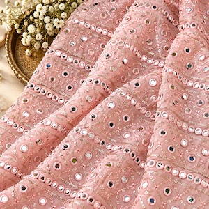 Peut inclure: Un tissu rose clair orné de broderies complexes et de petits miroirs circulaires disposés en bandes horizontales. Le textile présente un motif géométrique répétitif de lignes cousues et de paillettes, créant une surface texturée aux reflets brillants.