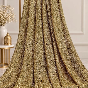 Könnte beinhalten: Ein Stoffstück, das mit kleinen, reflektierenden goldenen Pailletten bedeckt ist. Das Material ist über eine horizontale Stange drapiert und bildet vertikale Falten, die die dichte, schimmernde Textur und die metallisch goldene Farbe der Pailletten betonen.