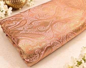 Puede incluir: Una pieza de tela doblada que presenta un color base rosa claro con un intrincado patrón de cachemira en oro metálico tejido. El diseño consiste en motivos repetitivos en forma de lágrima y detalles florales ornamentados en la superficie.