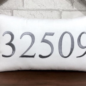 Könnte beinhalten: Weißes rechteckiges Zierkissen mit der gestickten Nummer "32509" in Grau. Das Kissen liegt auf einer dunklen Holzoberfläche, mit einer weißen Backsteinmauer im Hintergrund.