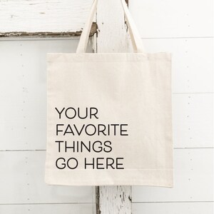 Op de afbeelding: Een cr&egrave;mekleurige draagtas met de tekst "YOUR FAVORITE THINGS GO HERE" in het zwart. De tas hangt aan een goudkleurige haak. De achtergrond is een witte, verweerde houten deur.