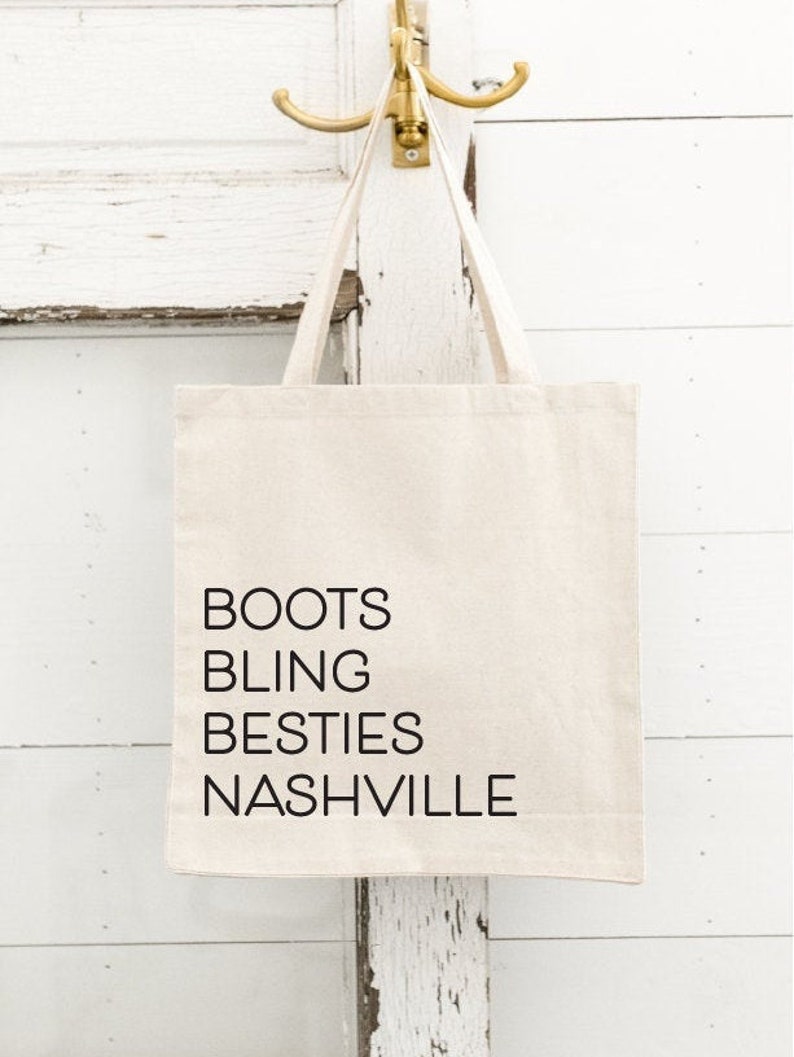 Op de afbeelding: Cr&egrave;mekleurige draagtas met zwarte tekst "BOOTS BLING BESTIES NASHVILLE". De tas hangt aan een goudkleurige haak aan een witte, verweerde houten deur.