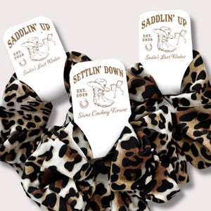 May include: Three scrunchies with a leopard print pattern. Each scrunchie has a white tag with the text "SADDLIN' UP" or "SETTLIN' DOWN", along with a cowboy boot, hat, and horseshoe graphic. The tags also read "Sadie's Last Rodeo" and "Same Cowboy Forever".