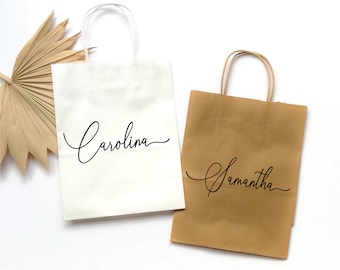 May include: Two paper gift bags with handles. The bag on the left is white with the name "Carolina" written in black cursive. The bag on the right is brown with the name "Samantha" written in black cursive.