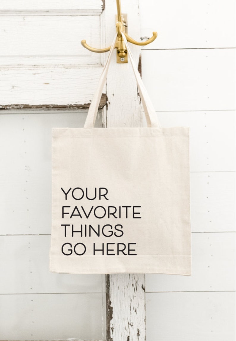 Op de afbeelding: Een cr&egrave;mekleurige draagtas met de tekst "YOUR FAVORITE THINGS GO HERE" in het zwart. De tas hangt aan een goudkleurige haak. De achtergrond is een witte, verweerde houten deur.
